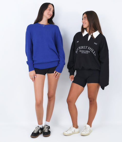 Two women wearing blue and black sweatshirts with visible branding on a white background